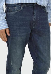 Dunkelblaue Jeans aus Denim mit klassischem Fünf-Taschen-Design, leichter Ausbleichung und geradem Bein. Hand auf dem Oberschenkel platziert.