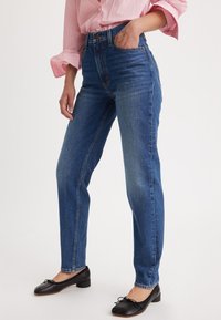 Jeans od plave trapero s visokim strukom i ravnim nogavicama. Imaju kopčanje na gumb, petlje za remen i blago zgužvane dijelove na bedrima.