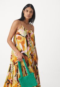 Yellow, patterned maxi dress with spaghetti straps and gathered neckline. Holding a small, woven green handbag.