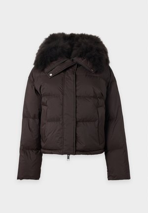 Vrouw draagt een donkerbruine donsjas met bontkraag, beige trui, zwarte wijde broekspijpen en zwarte laarzen met hak, staat met handen in de zakken.