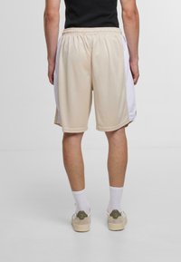 Beige Sportshorts mit weißen Seitenelementen, elastischem Bund und atmungsaktivem Material. Tragen mit weißen Socken und hellfarbenen Turnschuhen.