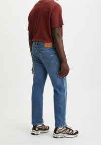 Vaqueros de mezclilla azul con un corte relajado, que presentan un parche de marca de cuero, un ligero desvanecimiento y dobladillos rematados, combinados con una camiseta burdeos y zapatillas.