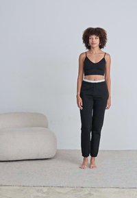 Top a bra cropped nero e pantaloni tapered neri con polsini elasticizzati e vita bianca. Tessuto morbido, modello in piedi su un tappeto chiaro.
