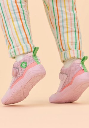 Kind met roze en groene sneakers en gestreepte broek, halverwege een stap tegen een beige achtergrond.