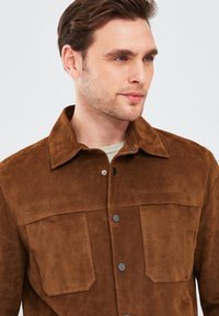 Bruine suède jas met een puntige kraag, twee borstzakken en zwarte drukknoopjes. Soepele textuur en een klassiek shirtjack ontwerp.