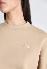 Sudadera beige con cuello redondo, que presenta un pequeño acento de logo texturizado. Tela suave con detalles de costura visibles.