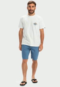 Witte katoenen T-shirt met een diamantgrafiek; blauwe shorts; zwarte slippers; staande pose voor een neutrale achtergrond.