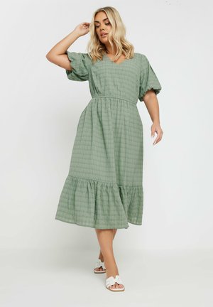 Vestido midi a cuadros verdes con mangas abullonadas, escote en V, cinturilla elástica y dobladillo con volantes. Llevado con sandalias blancas.
