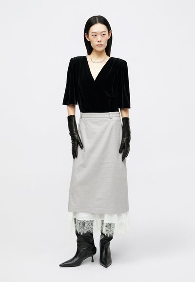 Γυναίκα με μαύρη βελούδινη μπλούζα, γκρίζα φούστα με άσπρη δαντελένια τρέσα, μαύρα δερμάτινα γάντια, μυτερές μαύρες μπότες και ασημένιο κολιέ, σε στάση.