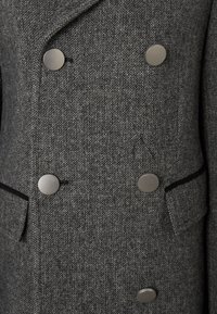 Šedý herringbone vlnený blazer s dvojitým zapínaním, šiestimi okrúhlymi striebornými gombíkmi a dvomi prednými vreckami s čiernym lemovaním.