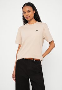 Mulher com cabelo encaracolado vestindo uma camisa bege de mangas curtas e calças pretas com um cinto castanho, contra um fundo branco simples.