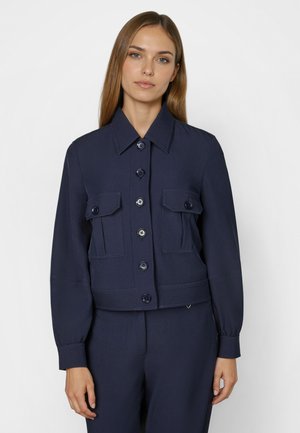 Frau mit glattem hellbraunem Haar trägt eine marineblaue, geknöpfte Jacke mit Klappentaschen und passende Hose, stehend vor einem schlichten Hintergrund.