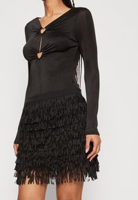 Frau in einem schwarzen Langarmkleid mit herzförmigen Ausschnitten an der Brust und einem geschichteten Fransenrock.