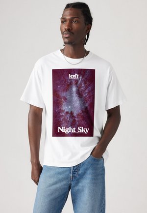 Muškarac s pletenom kosom nosi bijelu majicu s prikazom zvjezdanog neba kroz krošnje stabala, s natpisima "Levi's Explorer Division" i "Noćno nebo."