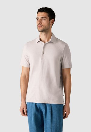 Mann, der ein hellbeiges kurzärmeliges Poloshirt und lockere blaue Hosen trägt, vor einem schlichten hellgrauen Hintergrund steht.