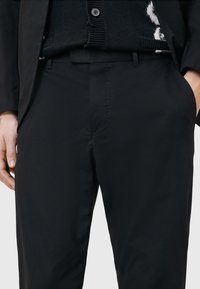 Zwarte broek van gladde stof, met een rechte snit, zijzakken en een subtiele knoopsluiting. Boven kleding een gestructureerd zwart trui.