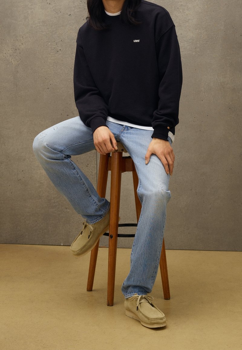 Felpa nera con il logo "LEVI'S", jeans chiari strappati, e scarpe beige slip-on, seduto su uno sgabello di legno contro uno sfondo neutro.