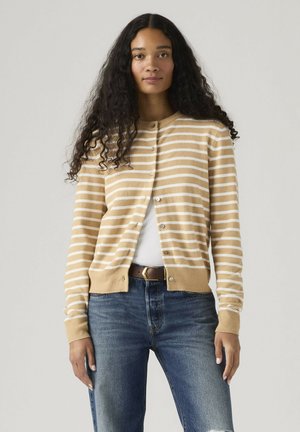 Kobieta o długich kręconych włosach, ubrana w beżowy, biało-paskowany kardigan na guziki, biały top, brązowy pasek i niebieskie dżinsy, stoi na tle jednolitego tła.