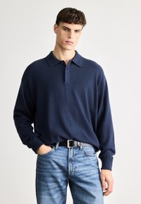 Ναυτικό μπλε πλεχτό με πόλο κολάρο και μακριά μανίκια, κατασκευασμένο από μαλακό, υφασμάτινο ύφασμα. Συνδυάζεται με ανοιχτό μπλε τζιν και μαύρη ζώνη.