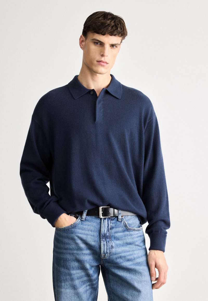 Ναυτικό μπλε πλεχτό με πόλο κολάρο και μακριά μανίκια, κατασκευασμένο από μαλακό, υφασμάτινο ύφασμα. Συνδυάζεται με ανοιχτό μπλε τζιν και μαύρη ζώνη.