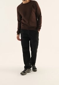 Sudadera marrón con cuello redondo, combinada con pantalones cargo negros con bolsillos laterales y zapatillas deportivas negras con acentos grises.