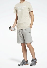 Beige Reebok t-shirt med stort vitt logotyp, grå träningsshorts med logodetalj, grå träningsskor med orange detaljer, och vattenflaska.