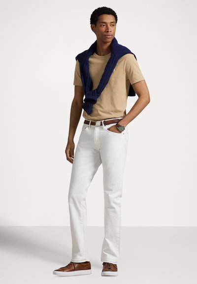 Λευκό κοντομάνικο μπλουζάκι, λευκό τζιν παντελόνι, καφέ ζώνη και καφέ αθλητικά παπούτσια. Μπλε σκούρο πουλόβερ κρέμεται στους ώμους. Ρολόι στον καρπό.