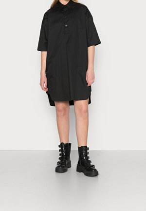 Schwarzes, übergroßes Hemd mit kurzen Ärmeln, einem Kragen und Seitenschlitzen. Kombiniert mit klobigen schwarzen Schnürstiefeln mit Riemen.