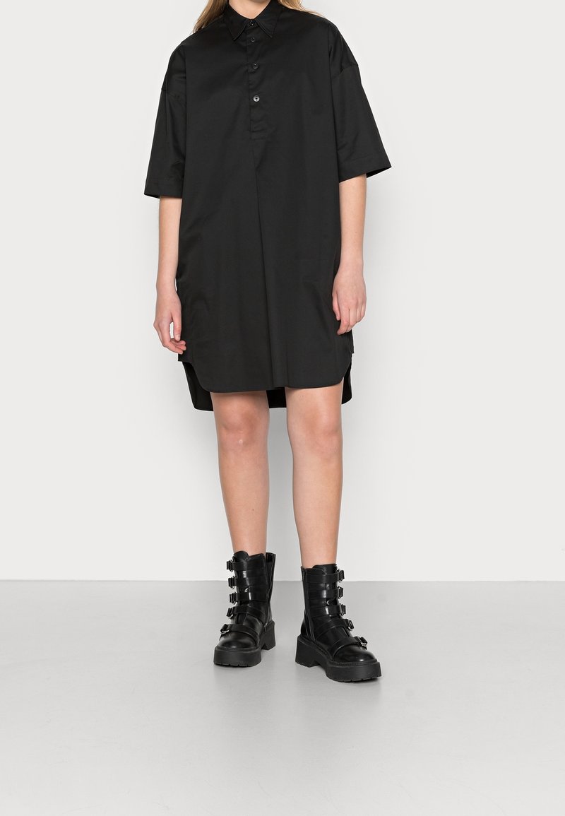 Chemise noire oversize à manches courtes, dotée d'un col et de fentes sur les côtés. Associée à des bottes montantes noires épaisses à lacets avec des sangles.