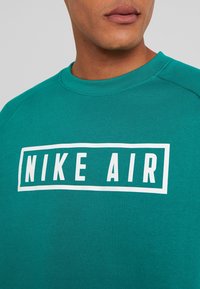 Turkos Nike sweatshirt med rund halsringning, som har en vit rektangulär logo med texten "NIKE AIR" som är tydligt placerad på bröstet.