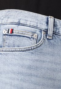 Los vaqueros de denim azul claro presentan un diseño clásico con un pasador para el cinturón, costuras visibles y una pequeña etiqueta roja, blanca y azul en el bolsillo.