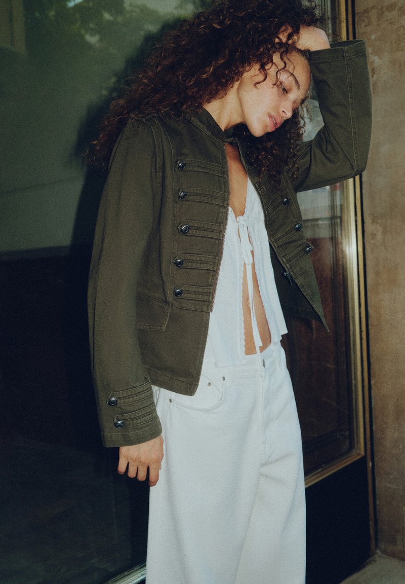 Femme aux cheveux bouclés portant une veste boutonnée vert olive par-dessus un crop top blanc noué et un pantalon blanc, debout à l’intérieur près d’une paroi en verre.