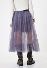 Lilla tylskørt med lagdelt tekstur, prydet med en elastisk talje. Båret med sorte ankelstøvler, der fremhæver et kontrasterende design.