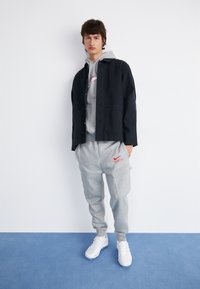 Chaqueta negra abotonada sobre un hoodie gris, combinada con pantalones de chándal grises con bolsillos y el logo rojo de Nike, y zapatillas deportivas blancas.