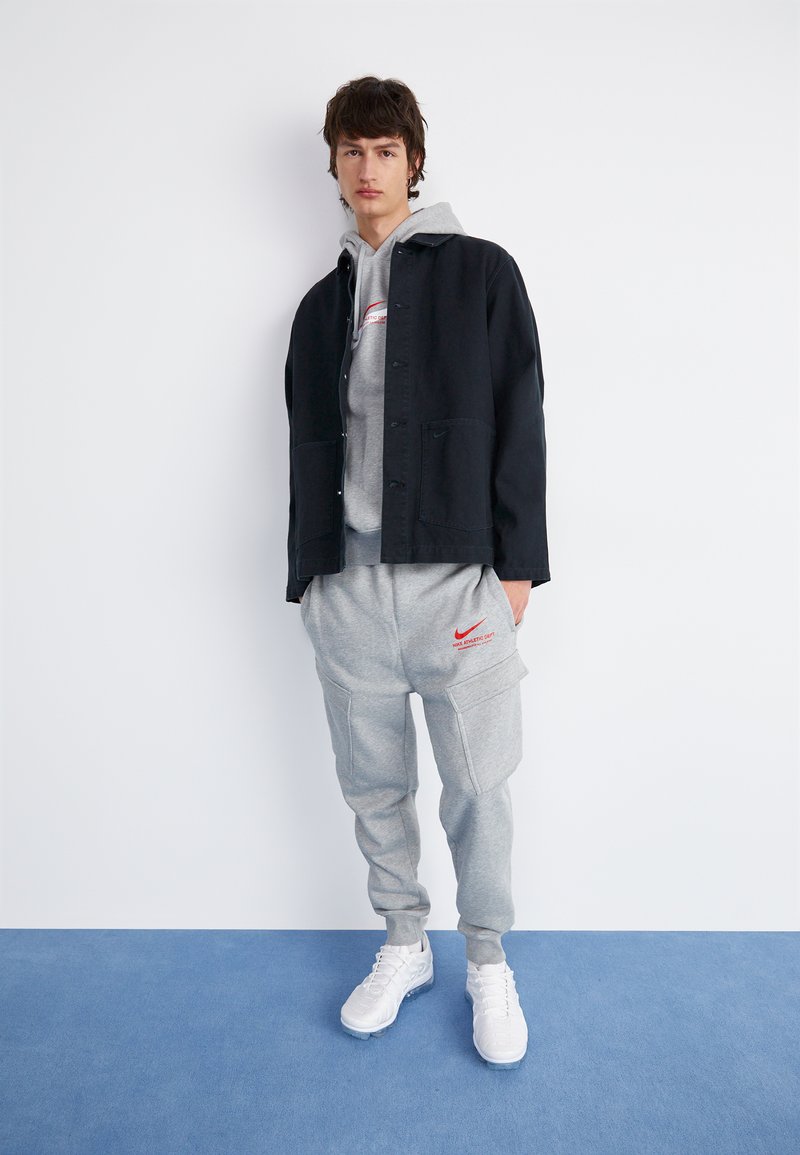 Chaqueta negra abotonada sobre un hoodie gris, combinada con pantalones de chándal grises con bolsillos y el logo rojo de Nike, y zapatillas deportivas blancas.