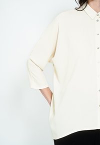 Hele beež värvi pluus kohandatud krae, nööbiga eest ja 3/4 varrukatega. Kangas on sile ja pehme tekstuuriga ning lõige on mugavalt lõdvestunud.