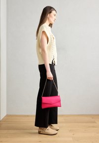 Žena stojící uvnitř, oblečená v krémovém pleteném bezrukávovém topu, černých širokých kalhotách, béžových dřevácích a držící jasně růžovou kabelku na zlatém řetízku.