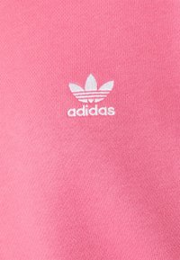 Vaaleanpunainen puuvillakangas, jolla on sileä pinta, esittelee valkoista Adidas-logoa, jossa on kolmonen muotoilu ja teksti alla, kiinteän taustan päällä.