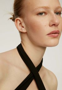Vestido negro de cuello halter que se cruza en el pecho, llevado con joyería mínima. El tono de piel parece claro, el cabello peinado en un moño. Fondo neutral.