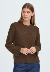 Brauner Strickpullover mit strukturiertem Muster, langen Ärmeln und geripptem Halsauschnitt. Kombiniert mit dunkelblauen Jeans, die einen lässigen Stil zeigen.