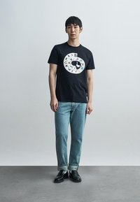 Μαύρο βαμβακερό t-shirt με κυκλικό λευκό σχέδιο και πολύχρωμα γραφικά. Συνδυάζεται με ανοιχτόχρωμα τζιν και μαύρα παπούτσια.