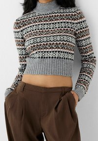 Grå korttröja med mönstrade detaljer i nyanser av brunt och svart, med hög halsringning och ribbat avslut. Matchas med bruna byxor.