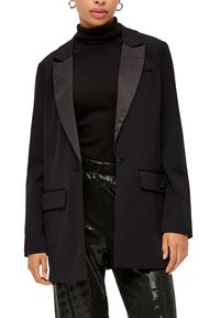 Black blazer with satin lapels, single button closure, and two front pockets. Worn over a black turtleneck and glossy sequined pants.