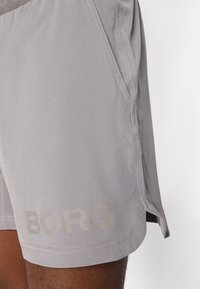 Hellgraue Sportshorts mit subtiler Glanzoberfläche, mit einem strukturierten Logo am Saum bedruckt, mit seitlichen Taschen und kontrastierenden Nähten.