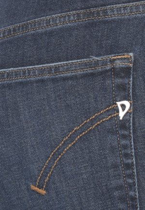 Primo piano della tasca posteriore di jeans in denim blu con cuciture marroni e un piccolo logo triangolare metallico sul bordo destro della tasca.