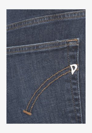 Primo piano della tasca posteriore di jeans in denim blu con cuciture marroni e un piccolo logo triangolare metallico sul bordo destro della tasca.