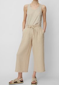 Beżowe lniane spodnie z szerokimi nogawkami, z elastycznym paskiem w talii i bocznymi kieszeniami, w połączeniu z jasnobeżowym topem na ramiączkach i sandałami na paski.
