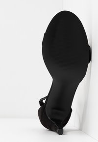 Chaussure à talon haut en daim noir avec un bout pointu et un talon aiguille. La semelle présente un motif texturé pour offrir adhérence et stabilité.