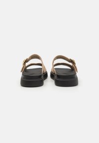 Tan leather sandals with black soles and adjustable ankle strap. Features a gold-tone buckle and an open back design.