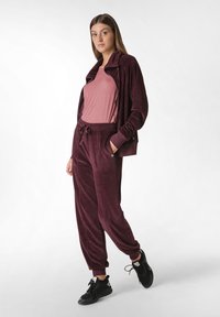 Bordeaux velours trainingspak bestaande uit een jasje met rits en broek met trekkoord. Het bovenstuk is gecombineerd met een lichtroze t-shirt en zwarte sneakers.